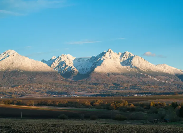 Poprad agricultural and forest lands with a panorama of snow-covered Tatra peaks under a blue sky.