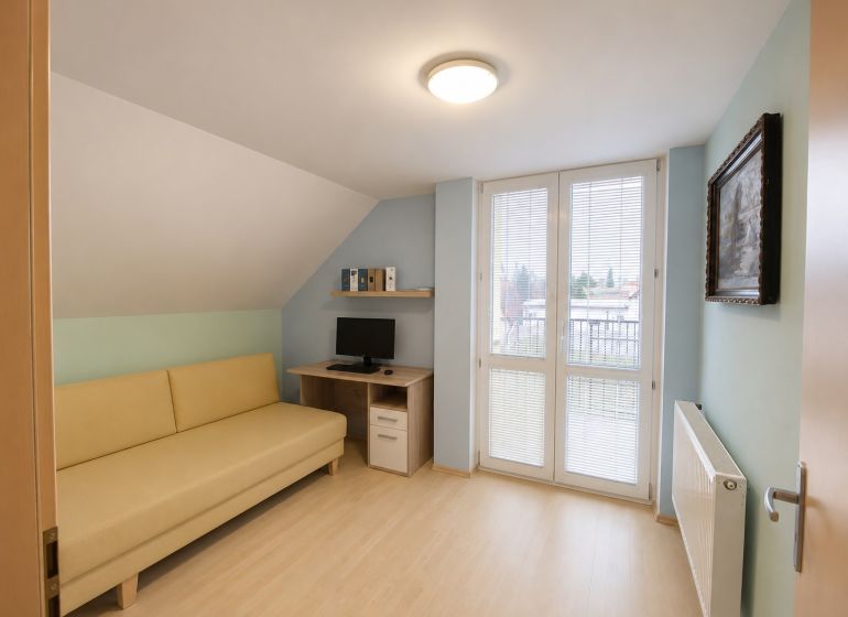 A room with a sofa, a computer desk, and a wooden-patterned floor in a family house.