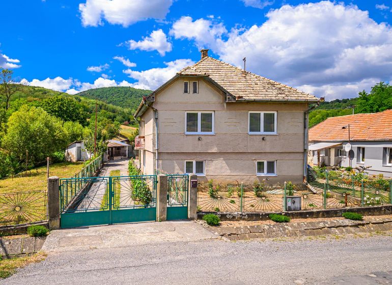 A family house in Hajnáčka surrounded by greenery and a fenced yard, with a blue sky.