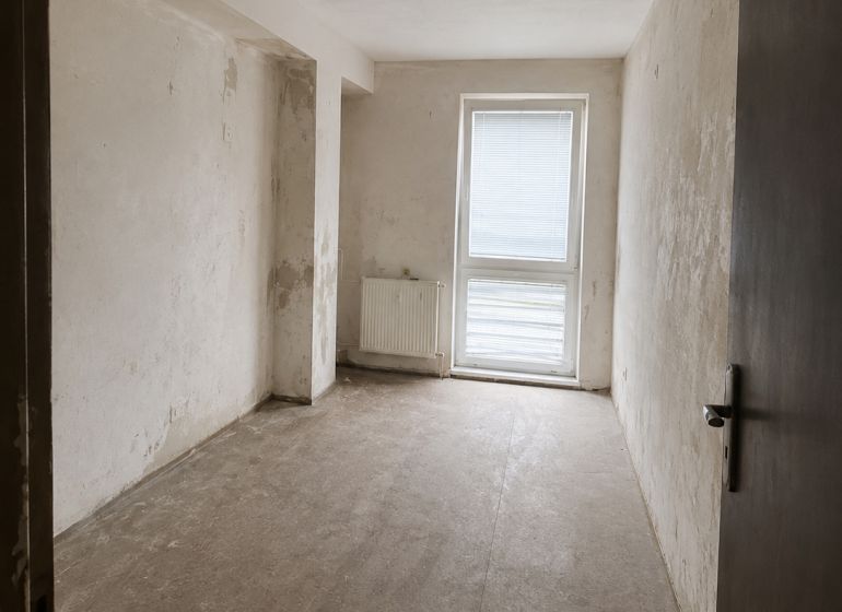 A view into an empty room in a two-room apartment with a concrete floor and balcony doors.
