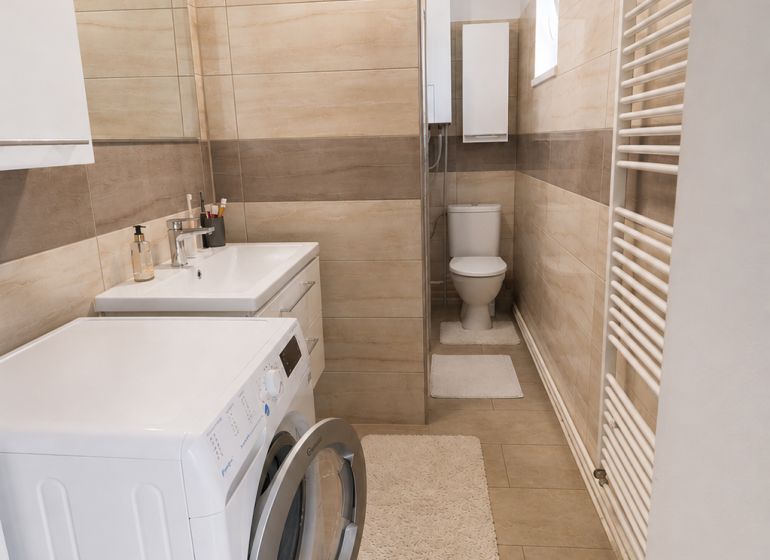 A bathroom in a family house with a washing machine, sink, shower, and toilet, with a wooden decor floor.