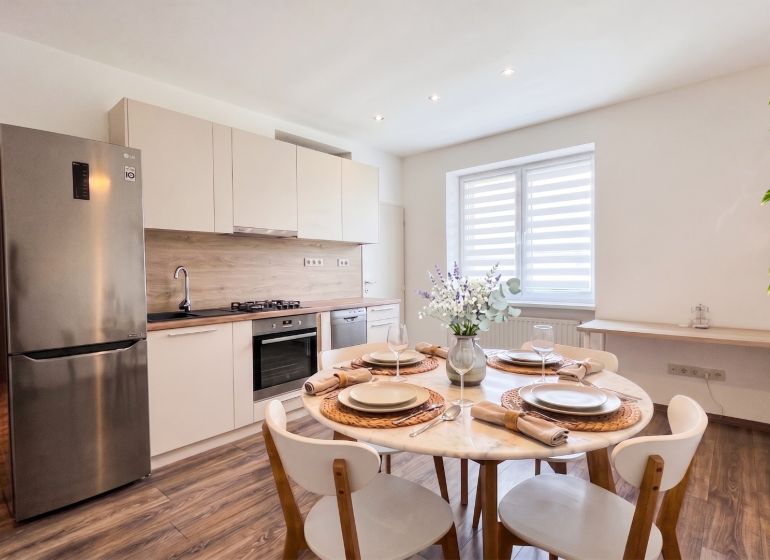 A kitchen of a two-room apartment with a wooden decor floor, a dining table, and appliances.