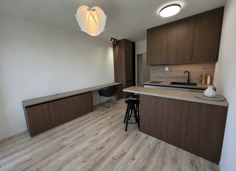 In the studio apartment, there is a kitchen unit with dark furniture and a floor with a wood design.