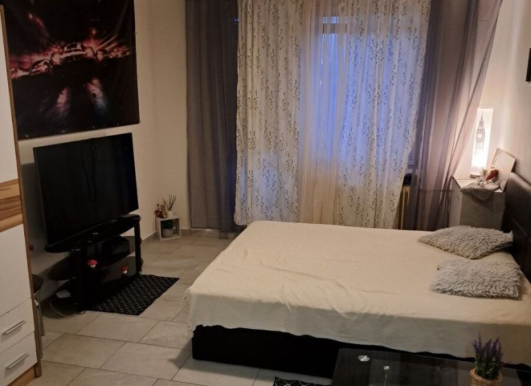 A studio apartment with gray-white walls, a bed, a television, and tiled flooring.