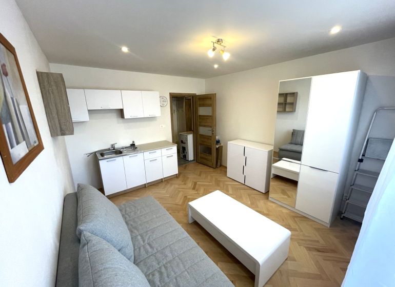 A studio apartment with a wooden decor floor, a kitchenette, and light-colored furniture.