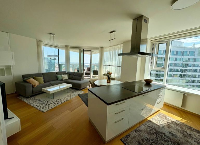 A bright living room with wood-patterned flooring in a three-room apartment, with a view from spacious windows.