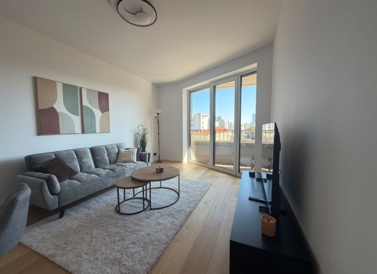Living room in a two-room apartment with a wooden decor floor, large window, and sofa.