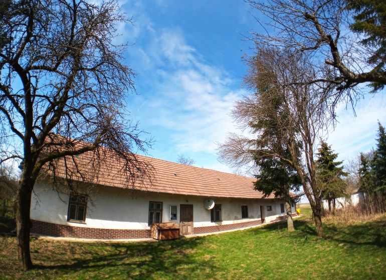 A family house in Buková, surrounded by trees, blue sky, white facade, tiled roof.