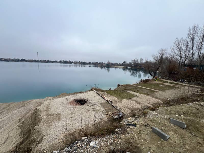 A view of a clear lake and natural surroundings at Veľké Košariská street. In the foreground, there are recreational plots with pebbles and a cold fire pit, while the surrounding landscape offers peace and quiet scenes typical of recreational areas.