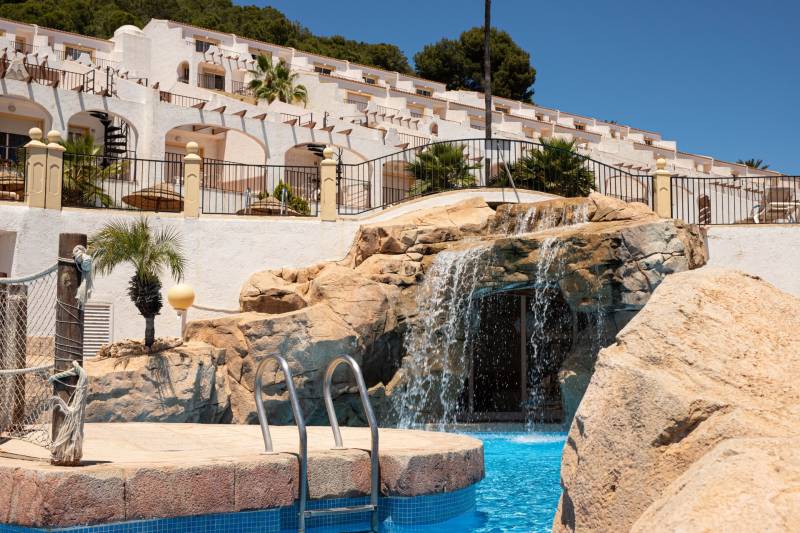 A waterfall by the pool in a recreational apartment in Calpe surrounded by palm trees and terraces.