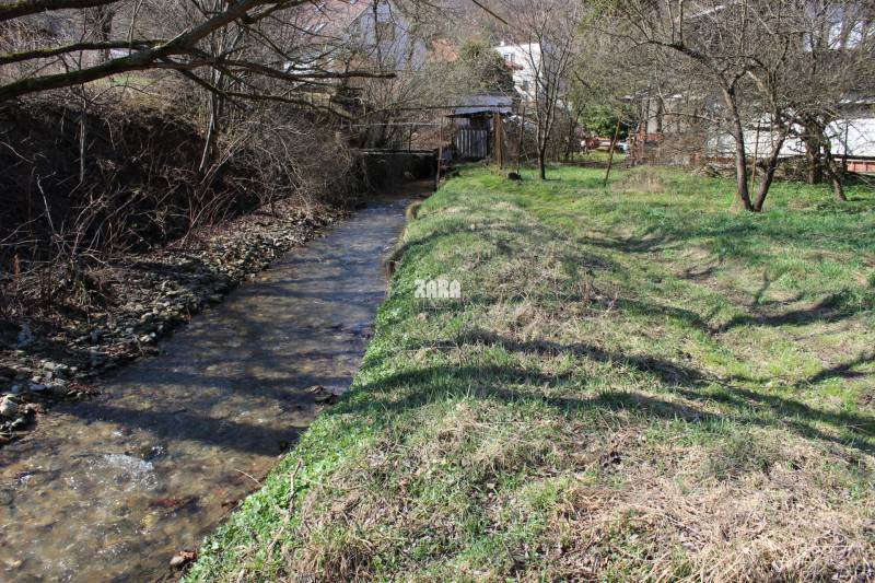 A stream with a grassy bank on Záhrady Street, surrounded by trees near a house, creates a peaceful view.