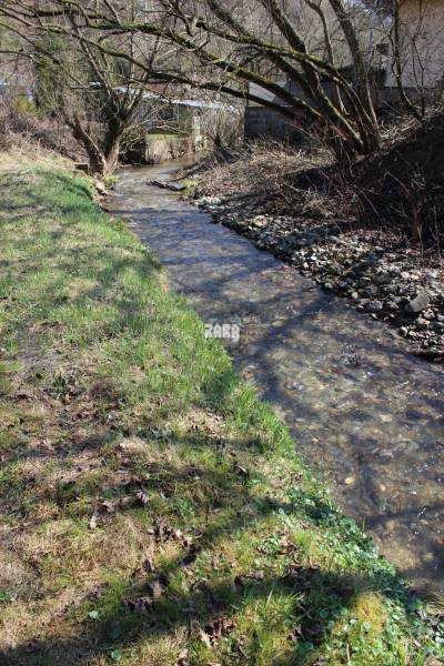 A stream surrounded by grass and trees, located on Myslavská and Záhrady streets.