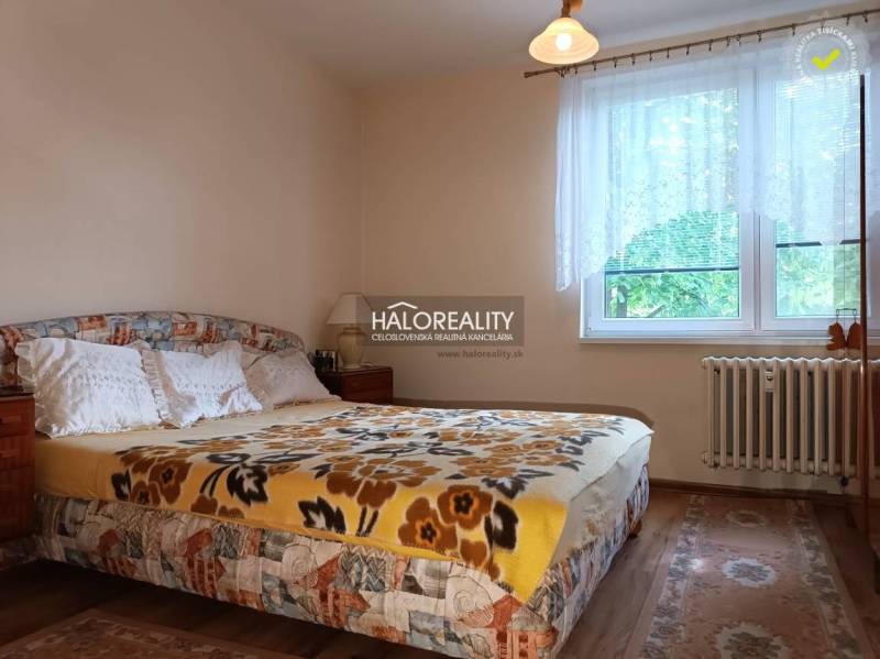 A bedroom in a 3-room apartment with a blanket, a lamp, and a window with white curtains.