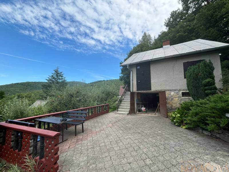 In the picture, there is a countryside cottage surrounded by nature with a terrace and a view of the forests.