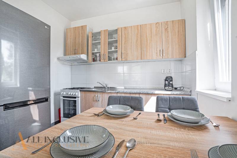 A kitchen in a 2-room apartment with a dining table, a stainless steel refrigerator, and a wooden kitchen unit.