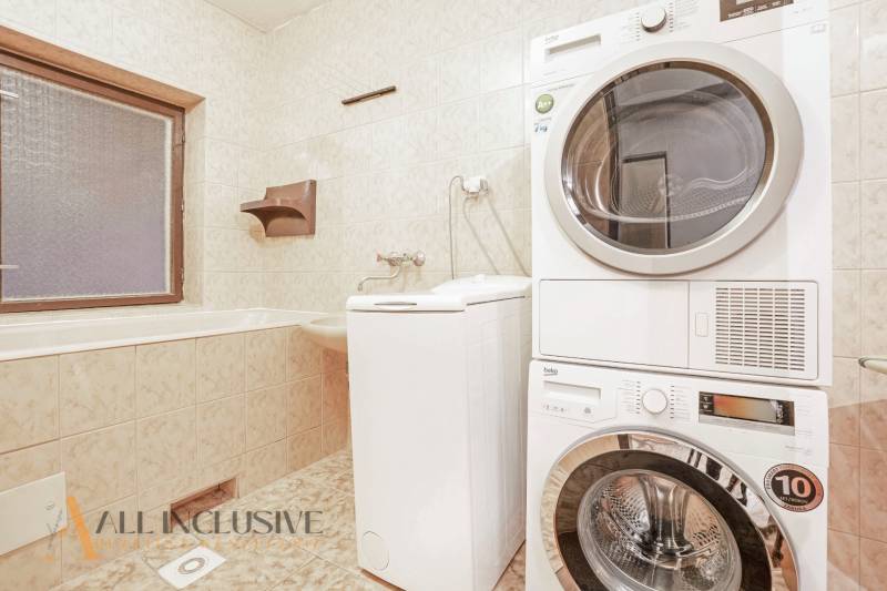 A bathroom in a family house with a window, a washing machine, and a dryer, cream tiles.