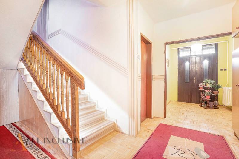 The interior of a family house with a wooden staircase, red carpets, and plants near the entrance.