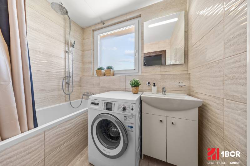 A bathroom with a bathtub, sink, and washing machine in a 3-room apartment full of light and plants.