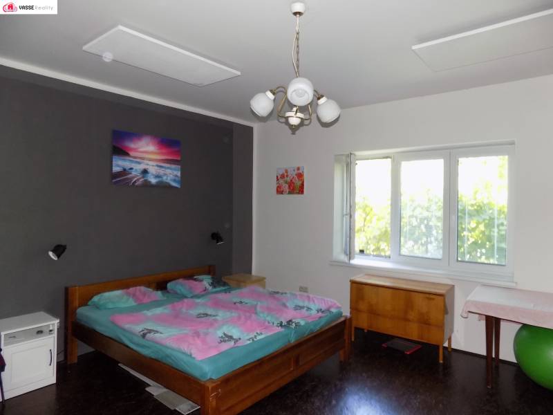 A bedroom in a family house with a dark wall, a bed, a dresser, and a large window.
