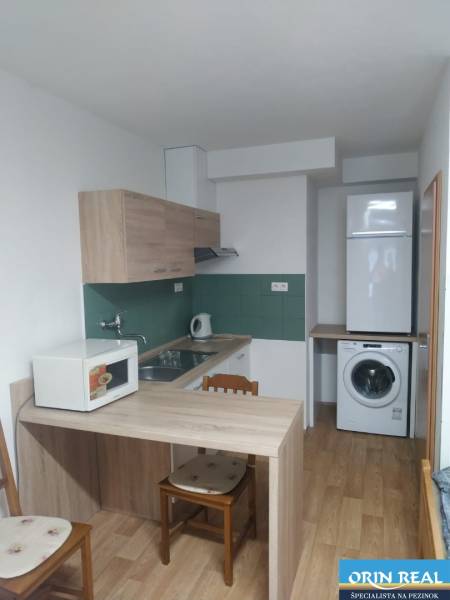 A compact kitchen with wooden furniture, a washing machine, and a microwave in a studio apartment.