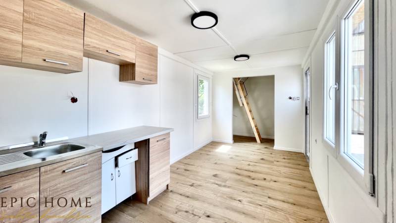 A kitchen in a cabin with a wooden floor, white walls, and a ladder to the upper floor.