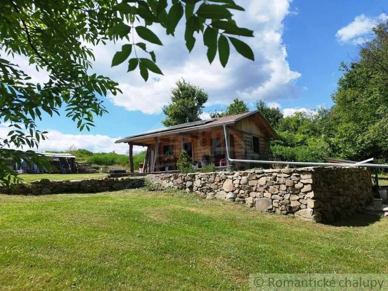 A cottage in Veľký Krtíš surrounded by nature, a stone wall, and a blue sky.