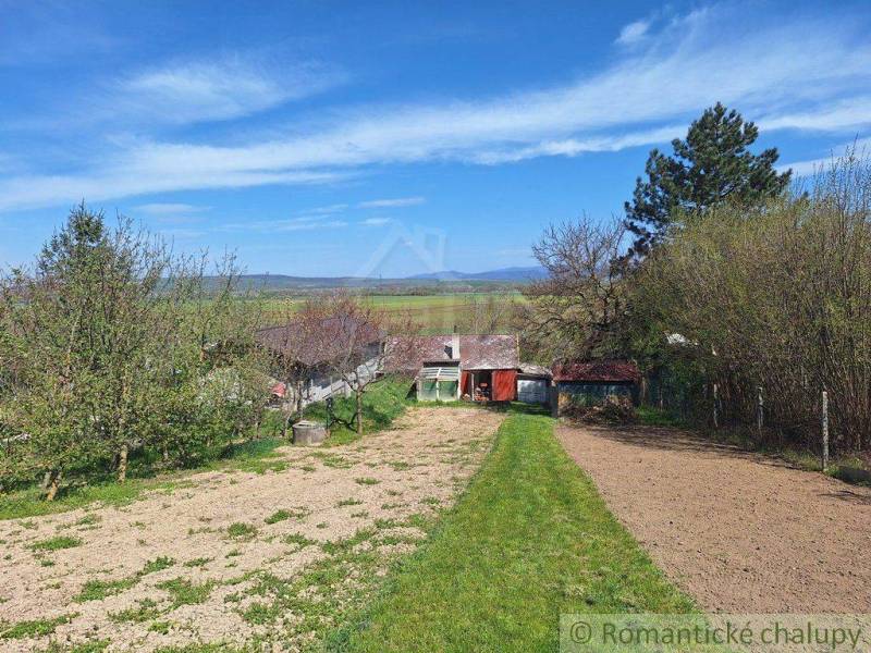 A cottage in the Nové Zámky area, surrounded by trees and a field under a blue sky.