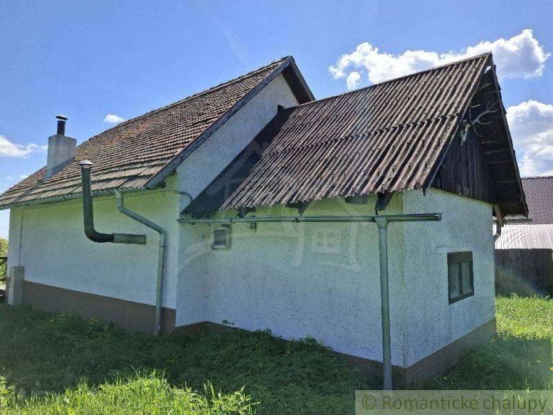 A cottage in Podkriváni with a tin roof and green surroundings under a blue sky.