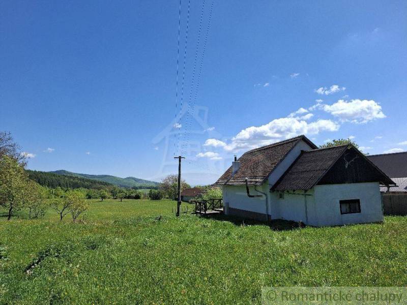 A cottage in Podkriváni surrounded by a green meadow and forests under a clear blue sky.