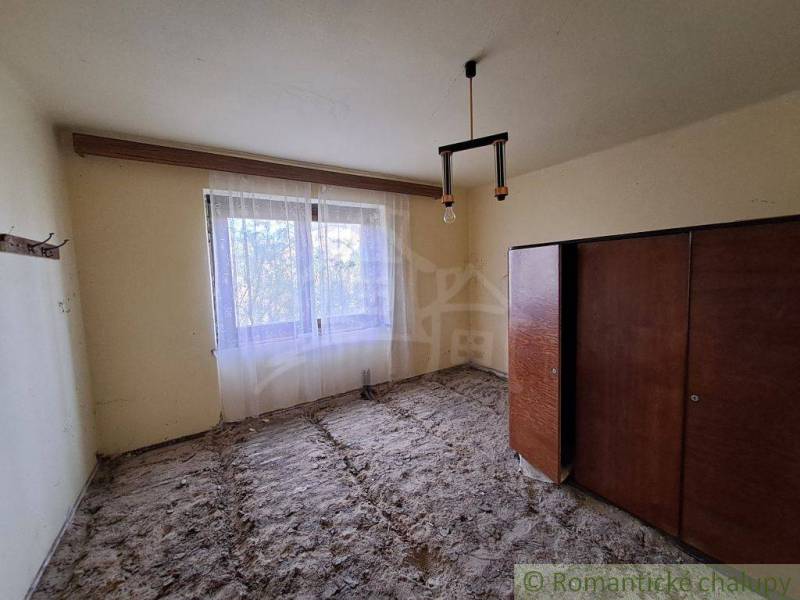 A room in a family house with a window curtain, old furniture, and an unfinished floor.