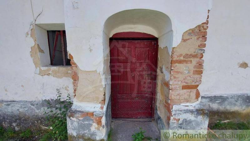 Old facade of a family house with peeling plaster, bricks, wooden door, window, greenery.