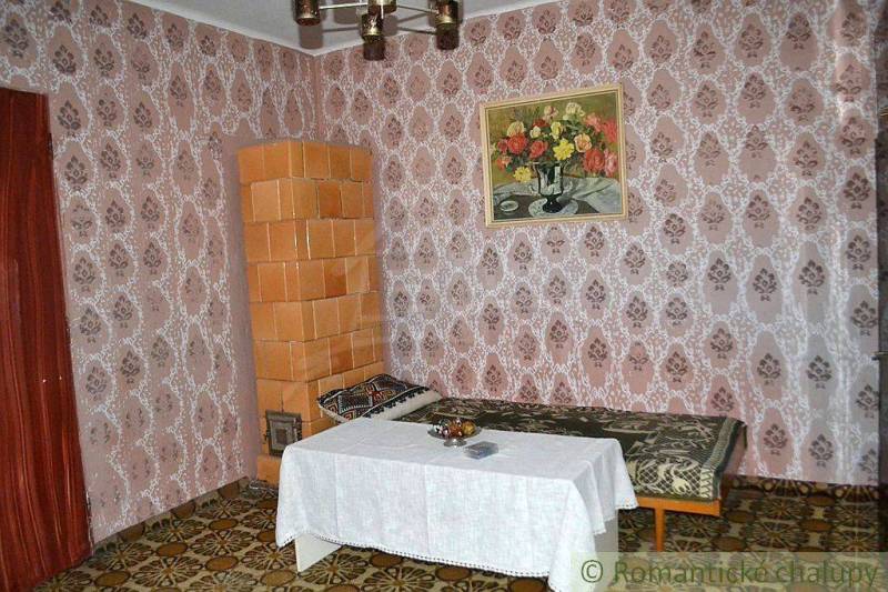 Interior of a family house: tiled stove, patterned wallpaper, sofa, table with tablecloth, painting of flowers.