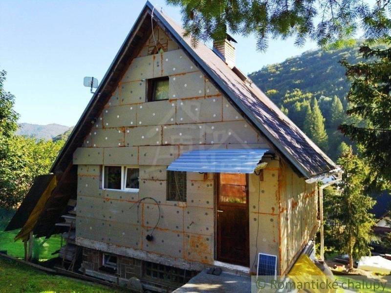 A cottage in a natural setting, sunlight illuminates the roof, surrounded by a garden and hills.