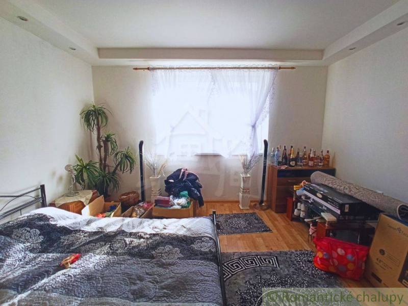 Bedroom in a family house, bed, plants, bottles, window with curtain, wooden floor.