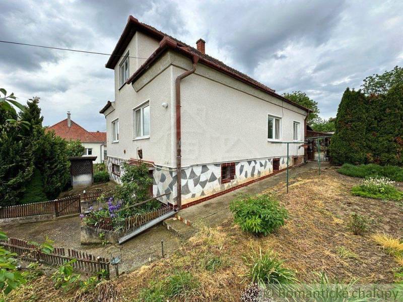 A family house with a white facade, garden, trimmed plants, fence, cloudy sky.