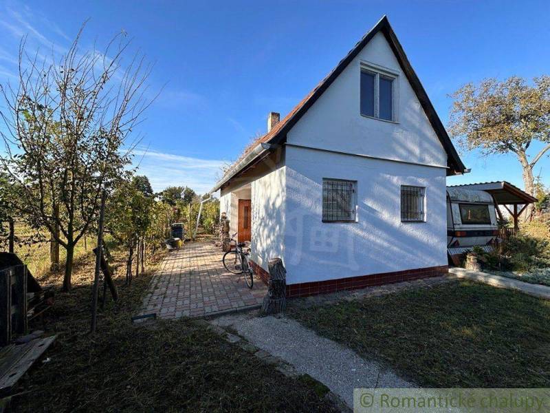 A simple white cottage with a bicycle, wooden door, trees, and a sunny background.
