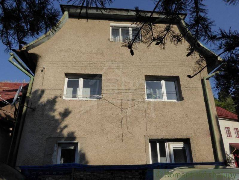 A family house with three windows, white plaster, and a shingle roof in a natural setting.