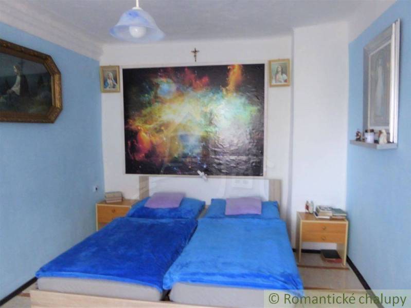 A bedroom with blue sheets, a colorful painting, nightstands, and pictures on the wall.