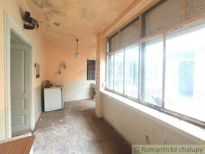 A veranda in an old cottage with large windows, a damaged ceiling, and a refrigerator.