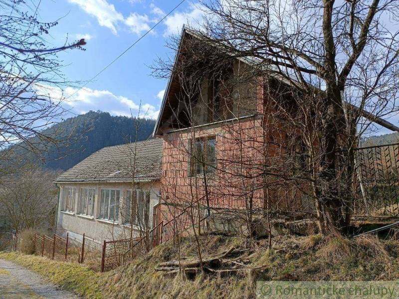 An old cottage in nature, surrounded by trees and a nearby mountainous environment.