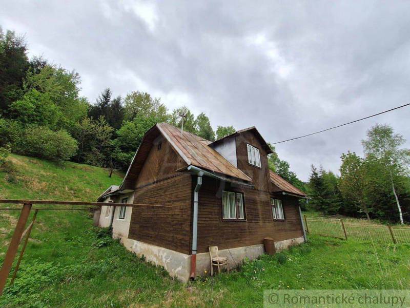 A wooden cottage in Veľké Rovné, surrounded by greenery and forests, under a cloudy sky.