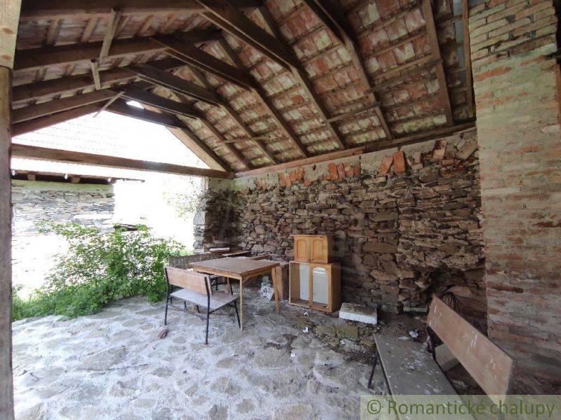 A stone stable with a wooden ceiling and simple furniture, surrounded by nature near a family house.