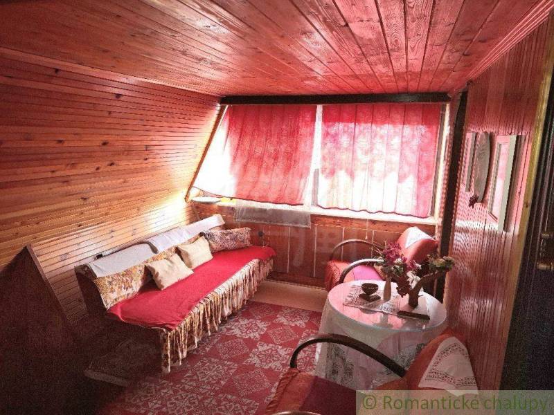 Cozy cabin interior, wooden paneling, red curtains, sofa, armchair, table, flowers.
