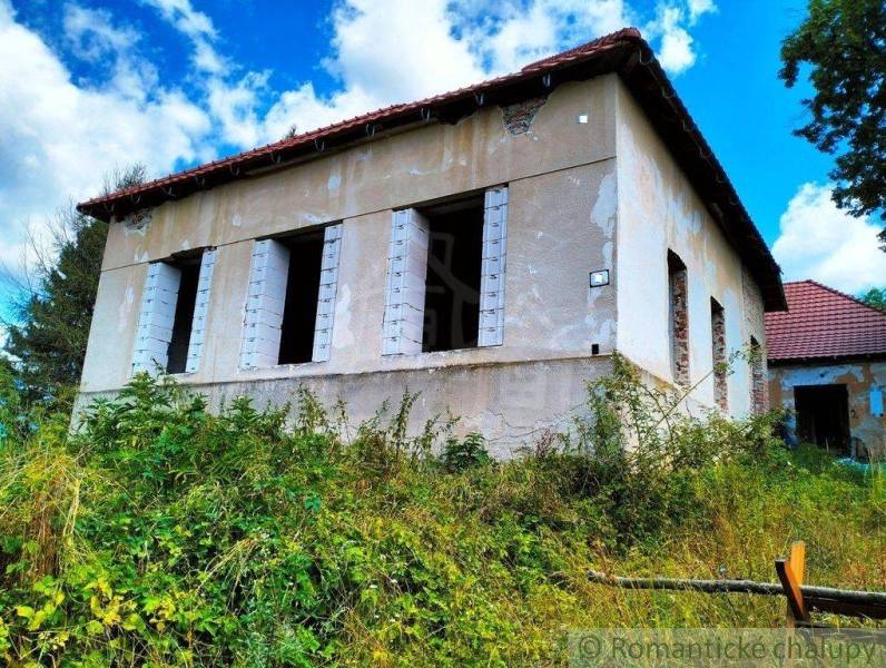 An abandoned family house with missing windows, surrounded by lush vegetation and a wooden fence.
