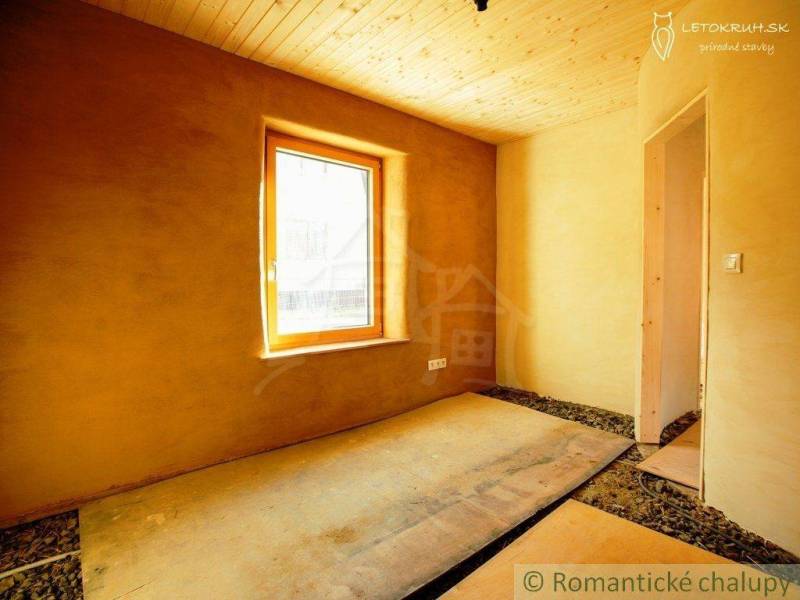 Interior of a room with a wooden ceiling, clay walls, a window, and a floor made of OSB boards.