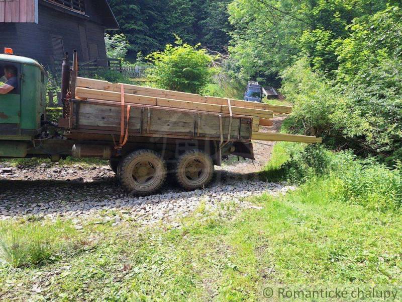 In the picture, there is a truck with wood by a cabin, surrounded by a forest and a path.