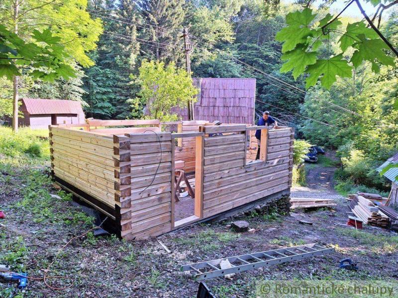 A wooden structure for a cabin is being built in the forest, surrounded by greenery and wooden cabins.