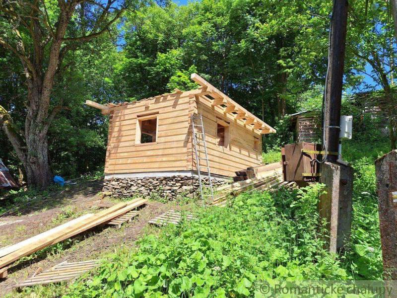 A wooden cabin on a construction site, surrounded by forest, with steep green terrain, ideal for relaxation.