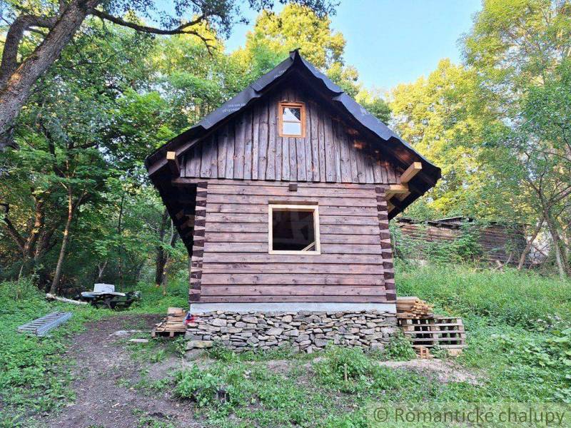 A wooden cottage in the middle of the forest surrounded by greenery and a wooden fence.