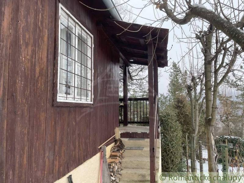 A wooden cabin with steps and firewood storage, with a snowy garden in the background.